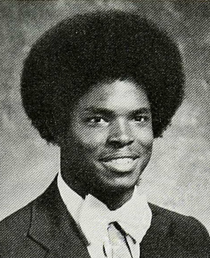 A Black teen is wearing a dinner jacket and white bow tie; with a large afro, he is facing right of the camera and smiling.